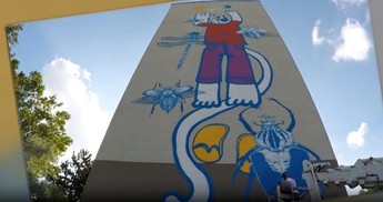 Das Foto zeigt eine Hauswand mit einem Graffiti, auf dem eine blaue Katze mit einer violetten Hose und einem roten T-Shirt zu sehen ist.