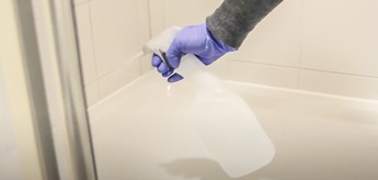 Man sieht eine Person, die die Innenseite einer Dusche reinigt, um die Entstehung von Schimmel zu verhindern. Sie trägt einen Handschuh und hält ein Reinigungsmittel in der Hand, mit dem die Wände der Dusche besprüht werden.