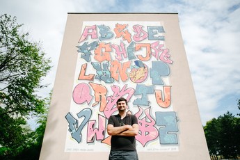 Foto eines Mannes in einem schwarzen T-Shirt vor einer Gemeindebauwand, die mit einem bunten Grafitti verschönert wurde. Das Grafitti zeigt ein abstraktes Alphabet mit Buchstabe in rosa, blau und orange. 