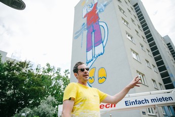 Foto eines Mannes in einem gelben T-Shirt vor einer Gemeindebauwand, die mit einem bunten Grafitti verschönert wurde. Das Grafitti zeig eine blaue Katze in buntem Gewand. 