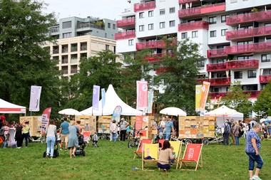Zahlreiche Besucher*innen beim Tag des Wiener Wohnbaus auf einer grünen Wiese vor modernen Wohngebäuden mit auffälligen roten Balkonen. Informationsstände, Liegestühle und Familien laden zum Verweilen ein.