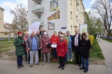 Das Foto zeigt eine Gruppe erwachsener Menschen in Winterjacken vor der Hauswand mit dem Wandmosaik.