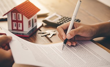 Ein Foto von einem Vertrag, der unterschrieben wird. Im Hintergrund sieht man ein kleines Haus, einen Schlüssel und einen Taschenrechner.