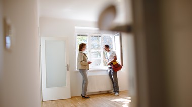 Zwei Menschen stehen in einer Wohnung vor einem Fenster und reden miteinander. Rechts im Bild sieht man eine Türklinke