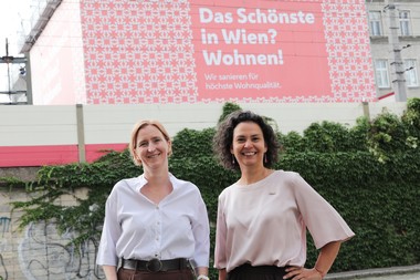 Elke Hanel-Torsch und Katharina Klement stehen vor einem Gebäude, mit Baustellentransparent. Darauf steht: Das Schönste in Wien? Wohnen!