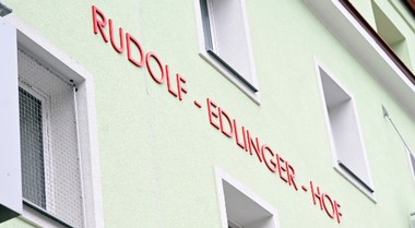 Das Foto zeigt eine hellgrüne Hausmauer mit Fenstern und dem roten Schriftzug "Rudolf-Edlinger-Hof".