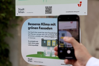 Foto einer Hand, die ein Handy im Kameramodus hält. Die Person scheint den QR Code einer Infotafel im Hintergrund öffnen zu wollen.