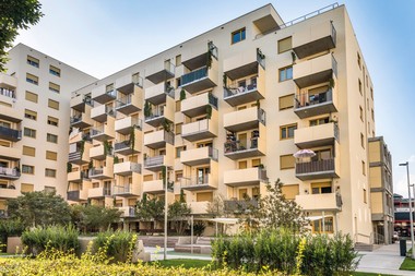 Innenhof des Gemeindebaus auf dem Areal des Eisring Süd. Das Gebäude hat teilweise eine orange/braune Fassade und die Stockwerke haben jeweils einen überdachten Balkon und im Erdgeschoss sind kleine Gärten