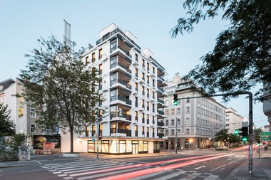 Ansicht des neuen Gemeindebaus an der Kreuzung Schönbrunner Straße und Reinprechtsdorfer Straße im 5. Bezirk. Das Gebäude ist weiß und es sind 7 Stockwerke mit Wohnungen zu sehen. An den ecken sind überdachte Balkone mit Netzen für Kletterpflanzen