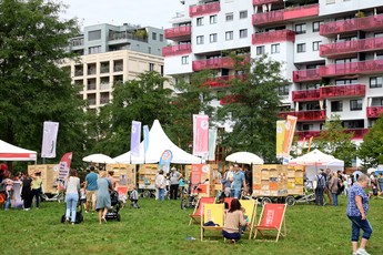 Zahlreiche Besucher*innen beim Tag des Wiener Wohnbaus auf einer grünen Wiese vor modernen Wohngebäuden mit auffälligen roten Balkonen. Informationsstände, Liegestühle und Familien laden zum Verweilen ein.