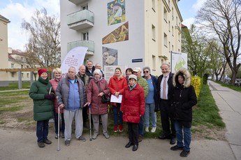 Das Foto zeigt eine Gruppe erwachsener Menschen in Winterjacken vor der Hauswand mit dem Wandmosaik.