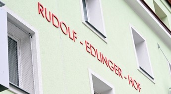 Das Foto zeigt eine hellgrüne Hausmauer mit Fenstern und dem roten Schriftzug "Rudolf-Edlinger-Hof".