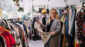 Foto einer freundlich lachenden Frau, welche zwischen langen Kleiderstangen steht und ein Oberteil in den Händen hält
