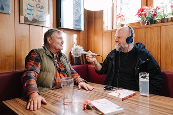 Zwei Personen sitzen in einem Café an einem Tisch; eine Person hält ein Mikrofon für ein Interview, beide lächeln. Bücher und Gläser liegen auf dem Tisch.