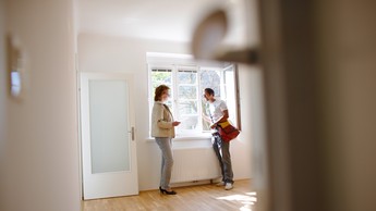 Zwei Menschen stehen in einer Wohnung am Fenster und reden. Rechts im Eck sieht man eine Türklinke.