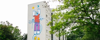 Foto einer grauen Hauswand, die mit einem bunten Graffiti verschönert wurde. Das Graffiti zeigt eine blaue Katze mit einem roten T-Shirt und einer violetten Hose. 