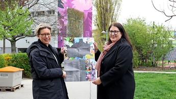 Wiener Wohnen Direktorin Karin Ramser und Wohnbaustadträtin Kathrin Gaal stehen vor einer mit violetten streetart verzierten Fassade eines Wohnhauses und halten einen Stadtplan in die Höhe, auf dem ein Kunstspaziergang eingezeichnet ist.  