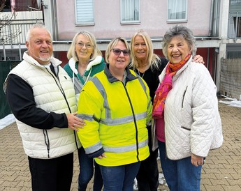 Magazincover „Mein Zuhause im Gemeindebau“ (Frühjahr 2026): Eine Frau mit Hut, Schal und beigem Mantel lehnt an einem Geländer vor einem Wohnhaus; Hinweis auf Gewinne und Titelstory „Zwischen Hinterhof und Hotelflur“.