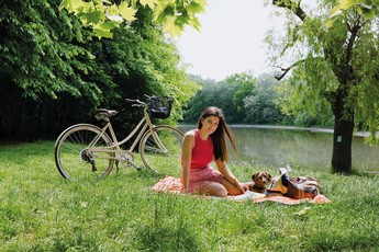 Foto von einer jungen Frau, die an einem Fluss auf einer Picknick-Decke sitzt. Neben ihr steht ein Fahrrad.