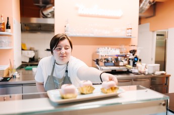 Eine Person hinter einer Ladentheke reicht ein kleines rosa Gebäck auf einem Teller nach vorne; im Hintergrund stehen eine Kaffeemaschine und Küchenutensilien.