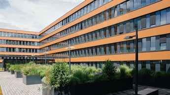 Moderner Bürokomplex mit orangefarbener Fassade und großen Glasfronten, umgeben von begrünten Hochbeeten im Innenhof. Ideal für Business-Standorte, nachhaltige Architektur und zeitgemäßes Arbeiten.