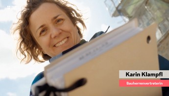 Eine fraue mit kurzen lockigen brauen Haaren lacht von oben in die Kamera. Im Vordergrund sieht man, dass sie eine Mappe mit Plänen hält.