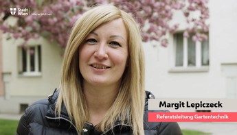 Eine blonde Frau lächelt ins Bild. Im Hintergrund sieht man eine Gebäudefassade und einen blühenden Kirschbaum.