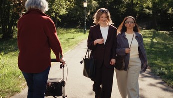 Eine alte Frau mit Rollator geht auf einem Weg im Park an zwei jüngeren Frauen vorbei.