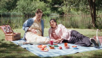 Eine schwangere Frau sitzt mit einer Freundin auf einer Picknickdecke auf einer Wiese, vor einem See.