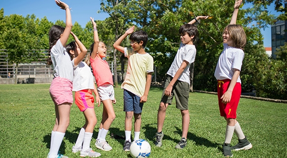Eine Gruppe von sechs Kindern steht auf einer grünen Wiese bei sonnigem Wetter. Die Kinder tragen sportliche Kleidung und Fußballschuhe. Sie haben Spaß, lachen und geben sich gegenseitig High-Fives. In der Mitte liegt ein Fußball auf dem Rasen. Im Hintergrund sind Bäume und ein Gebäude zu sehen – es wirkt wie ein Schulsportplatz oder ein Park.