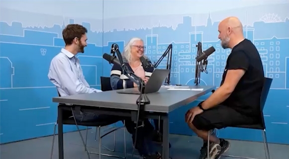 Drei Personen sitzen an einem Tisch in einem Podcast-Studio, jeweils mit einem Mikrofon vor sich. Auf dem Tisch steht ein Laptop. Im Hintergrund ist eine blau illustrierte Stadtlandschaft zu sehen.