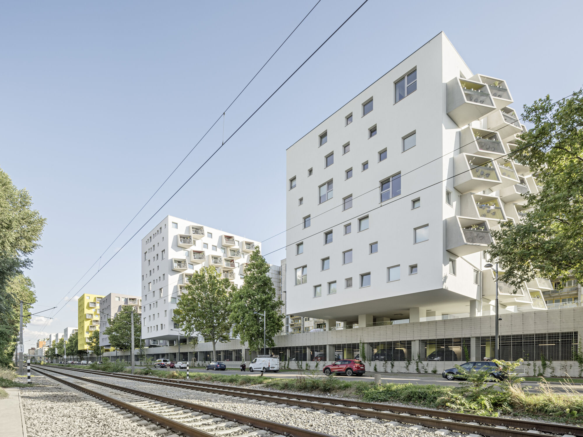 Das Bild zeigt das Rendering des Gemeindebau NEU am Handelskai 124A. Es ist ein weißes Gebäude direkt and er Straße mit bunten Balkonen.